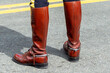 Rearview of high brown leather boots worn by a police officer with his dress uniform. The boots have a spur on the heels. The boots are worn by an adult on the pavement.