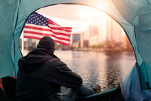 Homeless Man In A Tent In Focus. City And USA Flag Out Of Focus. Living In Hard Conditions Concept. Social Issue. Low Standard Of Living. Recession And Financial Crisis Effect On Population.