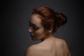 Pretty tan woman with ginger hair close her eyes and posing on a camera. The view from the back. Dark background