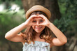 © _KUBE_ - Portrait of a little Caucasian girl in a straw hat showing a heart sign with her fingers. There is a park in the background. The concept of childhood, charity, peace and adoption of children