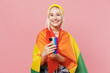 © ViDi Studio - Young blond lesbian woman 20s she wrapped in flag wear colorful knitted top yellow hat hold in hand use mobile cell phone isolated on plain pastel light pink background. People lgbtq lifestyle concept