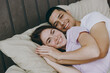 © ViDi Studio - Young smiling happy fun calm lovely caucasian couple two family man woman she he wearing t-shirt pajama lying in bed rest spending time together in bedroom home in own room hotel. Real estate concept.