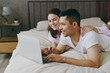© ViDi Studio - Side view young couple two family man woman she he wear t-shirt pajama hold use work on laptop pc computer lying in bed rest spend time together in bedroom home in own room hotel. Real estate concept.