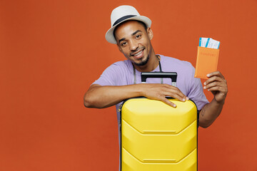 Traveler black fun happy man wear purple t-shirt hat hold suitcase passport look camera isolated on plain orange background Tourist travel abroad in spare time getaway Air flight trip journey concept.