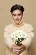 © Tatiana - A handsome young man with fair skin with dark, short, beautiful hair combed back in a beige turtleneck with a high neck stands on a dark beige background with a bouquet of daisies.Vertical studio shot