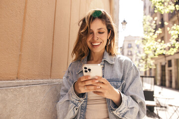 Smiling young caucasian woman holding smartphone smiling standing on city street. Brown-haired girl with wavy hair wears denim jacket. Online entertainment concept