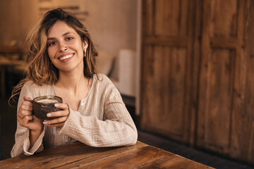 Positive young caucasian woman drinking hot coffee sitting in spacious cafe. Blonde smiles looking at camera, wears shirt. Concept of enjoying weekend, vacation