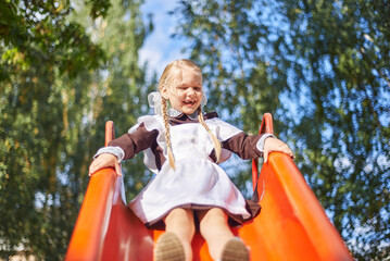 little girl schoolgirl plays in uniform in the yard. The child rolls from the children's slide on the playground.