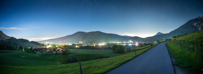 Poster - Panoramic shot of a road nearby a green field and city lights with a night sky behind the mountains