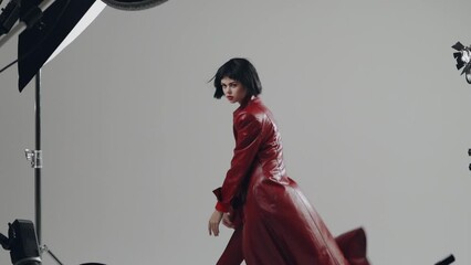 Wall Mural - Woman professional fashion model posing in a stylish red coat in the studio for a photo on a white cyclorama with studio fixtures. Posing like a dance