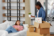 © Syda Productions - moving, people and real estate concept - happy smiling woman unpacking box and her friend resting on sofa at new home