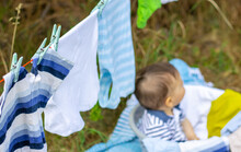 Cute Little Baby Boy Sit Outside In Garden On Picnic Blanket Laundry Drying On Rope In The Back.adorable Boy Rubs With Hands His Eye,sleepy Toddler.housekeeping Maternity.kid Clothes With Clothes Pin.