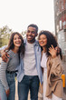 Three young interracial students with good mood are standing in embrace outdoors. Brunettes guy and girls wear casual clothes. Happy day concept