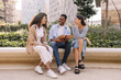 Three nice young interracial guys and girls are having nice conversation and discussing their homework sitting on street. Brunette students wear casual clothes in spring. Communication concept