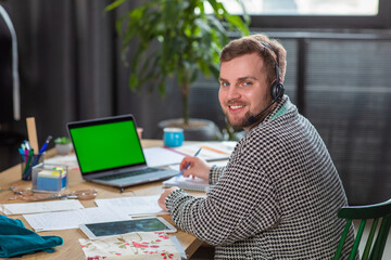 In front of the camera at tailoring atelier the tailor man have a online conversation with customer he make some notes on the notebook the desktop of laptop are green