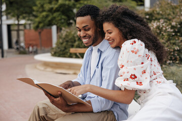 Wall Mural - Smiling young african classmates check their homework before class sitting outside. Guy wears shirt, girl is brunette blouse. Education concept