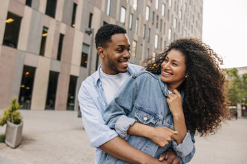 Wall Mural - Beautiful young african couple looking at each other hugging standing outdoors. Brunette guy and girl wear casual clothes. Concept of enjoyment