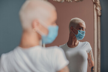 Wall Mural - Millenial young woman in medical protective face mask with short blonde hair portrait standing at mirror indoor