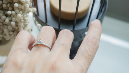 Wall Mural - Close up of an elegant diamond ring on woman finger with white flower, sunlight and shadow background. love and wedding concept. Soft and selective focus.