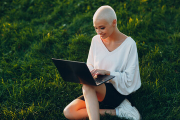 Portrait of young smiling millenial european short haired woman using laptop at green grass meadow in park. Beautiful happy blonde girl outdoor. Summer fashion female clothing.