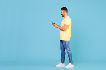 Full body young smiling happy caucasian man holding mobile phone in hand isolated on blue background studio portrait. People lifestyle concept