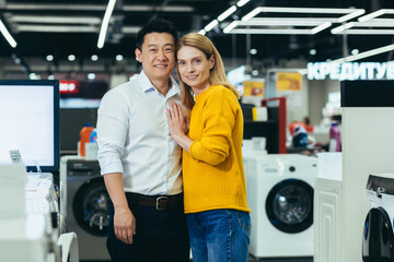 Wall Mural - Portrait of a young married couple shopping in a home appliance supermarket, a diverse family of a man and a woman smiling and looking at the camera, choosing and buying home appliances