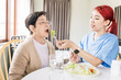 © artitwpd - Asian woman caregiver wearing medical scrub takes care of senior Asian woman by feeding vegetarian salad to eat at home. caregivers visit home Home health care concept and nursing home.