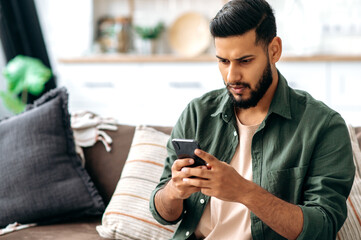 Puzzled indian or arabian guy in casual wear, sits on sofa in a living room, using smartphone, texting online, browsing social networks, reads news, looks at screen in confusion. Wireless technologies