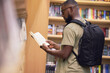 © Sanele Gobinduku/peopleimages.com - Education student with book in a library at university, college or school reading or doing research. Scholarship, learning and study knowledge and black man with print books on shelf and backpack