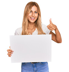 Poster - Beautiful blonde young woman holding blank empty banner smiling happy and positive, thumb up doing excellent and approval sign