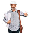Middle age grey-haired man wearing safety helmet holding blueprints smiling happy and positive, thumb up doing excellent and approval sign