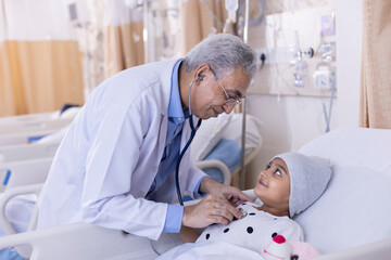 Doctor examining little girl patient lying on bed by stethoscope at hospital
