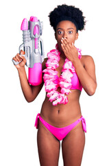 Poster - Young african american woman wearing bikini and hawaiian lei holding water gun covering mouth with hand, shocked and afraid for mistake. surprised expression