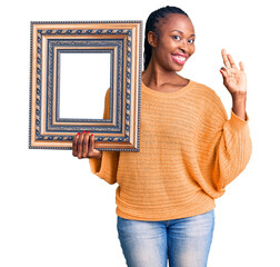 Wall Mural - Young african american woman holding empty frame doing ok sign with fingers, smiling friendly gesturing excellent symbol