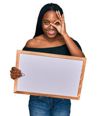 Wall Mural - Young african american woman holding empty white chalkboard smiling happy doing ok sign with hand on eye looking through fingers