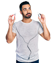 Poster - Young hispanic man with beard holding battery clamps smiling looking to the side and staring away thinking.