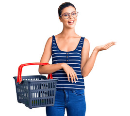 Sticker - Young beautiful woman holding supermarket shopping basket celebrating victory with happy smile and winner expression with raised hands