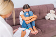 unhappy crying pre-adolescent girl talks to psychologist, hugging her knees at home or in office, expressing emotions. sad teenage girl listens to psychologist at meeting, thinking about her problems.