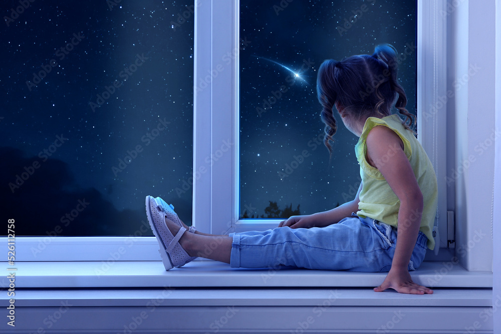 Cute little girl sitting near window and looking at shooting star in ...