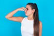 beautiful brunette woman wearing white tank top over blue background smelling something stinky and disgusting, intolerable smell, holding breath with fingers on nose. Bad smell