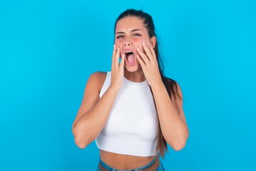 Wall Mural - Stupefied beautiful brunette woman wearing white tank top over blue background expresses excitement and thrill, keeps jaw dropped, hands on cheeks, has eyes popped out