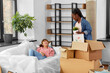 © Syda Productions - moving, people and real estate concept - happy smiling woman unpacking box and her friend resting on sofa at new home
