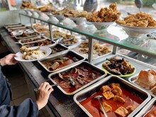 Padang Food Stalls Share A Variety Of Buffet Menus With Vegetables, Chicken Curry Sauce, Beef, Eggs, Tofu, Tempeh. The Waiter Prepares The Food. Menu In Glass Display Case. Asia And Asian Food. Pile.