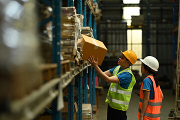 Two storehouse employees wearing hard hats and reflective jackets loading or unloading boxes on the shelf in warehouse