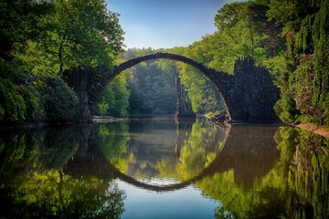 Wall Mural - Beautiful view of an arched bridge in the forest symmetrically reflecting on the river water