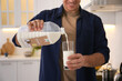 Man pouring coconut milk from gallon bottle into glass in kitchen, closeup. Vegan product