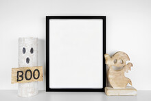 Halloween Mock Up. Black Frame On A White Shelf With Rustic Wood Ghost Decor. Portrait Frame Against A White Wall. Copy Space.