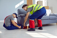 Loader Gets Injured At Work. Man Who Got Leg Caught Under Sofa Holding His Broken Leg With Expression Of Pain On Face While The Other Guy Is Trying To Help And Lift The Sofa. Accident, Injury Concept