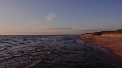 Wall Mural - Panoramic view of a deserted beach washed by sea waves, turning into a view of the sea distance with the rising sun reflected in the sea water. Video from a drone.