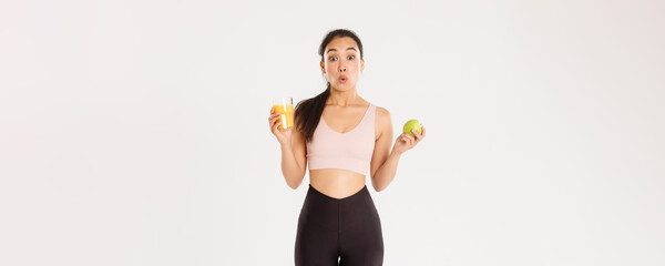 Sport, wellbeing and active lifestyle concept. Amused and wondered cute asian girl likes fitness and eating healthy food, holding apple and orange juice, looking amazed saying wow, white background